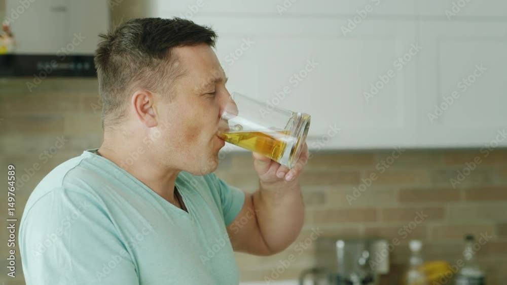 A young Caucasian man with pleasure drinks beer in his kitchen. Then he smiles and looks into the camera. Portrait of a lover of beer A young Caucasian man with pleasure drinks beer in his kitchen. Then he smiles and looks into the camera. Portrait of a lover of beer