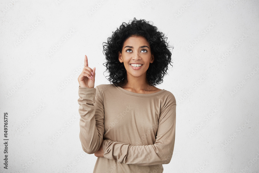 Waist up shot of joyful girl wearing beige long sleeve t-shirt looking up, pointing finger at copy space above her head. Black young woman indicating something on blank studio wall with hand - obrazy, fototapety, plakaty 