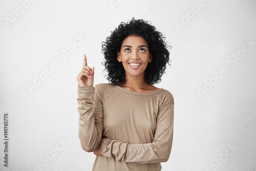 Waist up shot of joyful girl wearing beige long sleeve t-shirt looking up, pointing finger at copy space above her head. Black young woman indicating something on blank studio wall with hand
