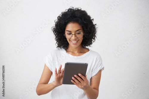 Studio shot of dark-skinned smiling charismatic and beautiful female student holding modern gadget, using tablet for video call with her friends, watching funny videos or doing homework, chatting