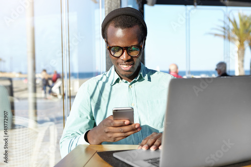 Handsome Afro American student wearing stylish accessories typing message on mobile phone, sitting at cafe in front of laptop pc, waiting for friend for lunch, using free wi-fi internet connection