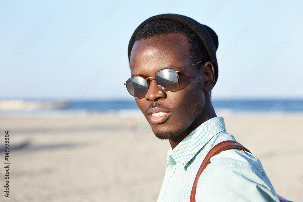 People, fashion, lifestyle and travel concept. Outdoor summer shot of fashionable young Afro American man in headwear and eyeglasses spending vacations by the ocean, contemplating amazing view People, fashion, lifestyle and travel concept. Outdoor summer shot of fashionable young Afro American man in headwear and eyeglasses spending vacations by the ocean, contemplating amazing view