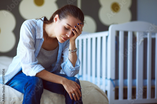 Young tired woman sitting on the bed near childrens cot