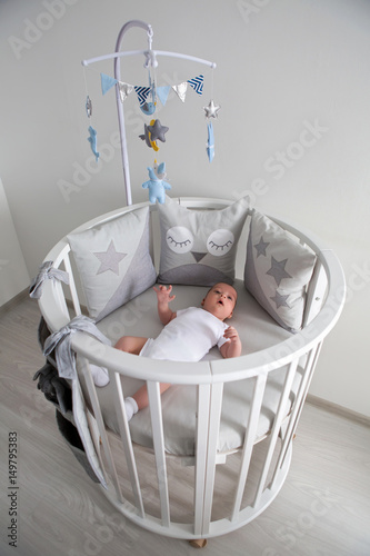 newborn lies in the round white bed with mobile and toys