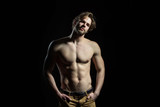 Young man holds his hands in his pockets. Attractive handsome man on black background with naked torso and sporty muscles. Sexy guy