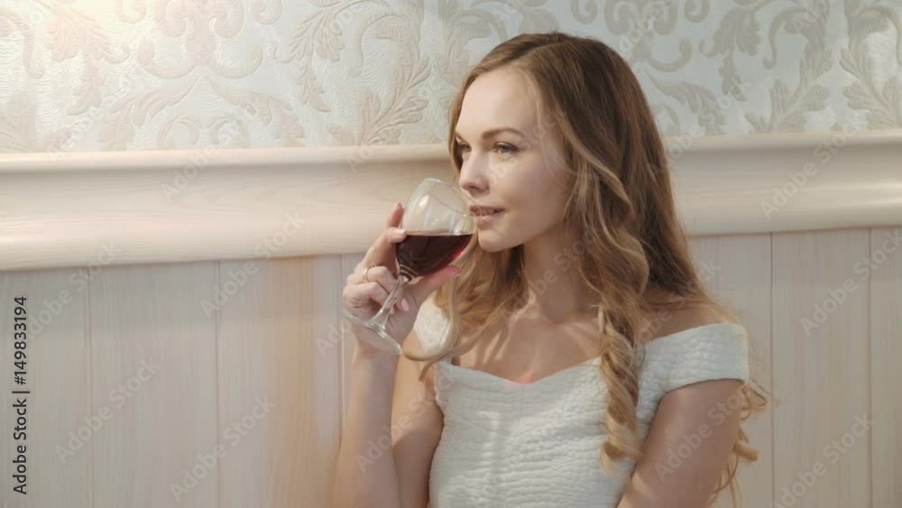 Young business woman relaxing on the bed Drinks from a glass of red wine at her hotel bedroom. on bed in luxurious room interior. Young beautiful girl in white dress Young business woman relaxing on the bed Drinks from a glass of red wine at her hotel bedroom. on bed in luxurious room interior. Young beautiful girl in white dress