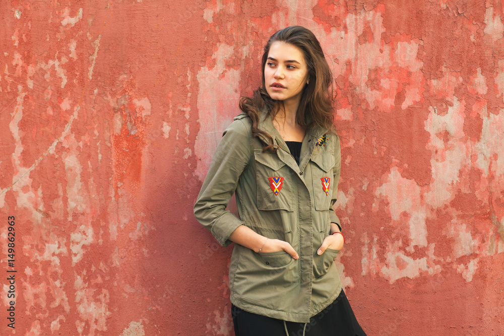 young female looks left in military style jacket near red dilapidated concrete wall young female looks left in military style jacket near red dilapidated concrete wall
