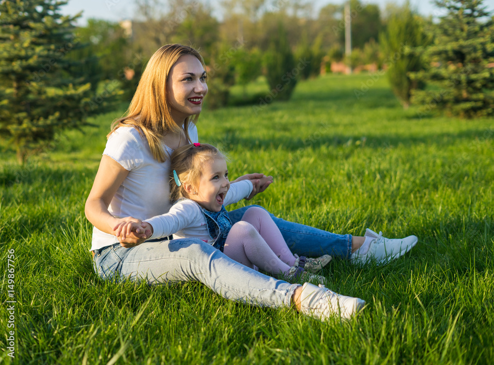 Portrait of smiling beautiful young woman and her small daughter sitting on green lawn on grass in summer park. Portrait of smiling beautiful young woman and her small daughter sitting on green lawn on grass in summer park.