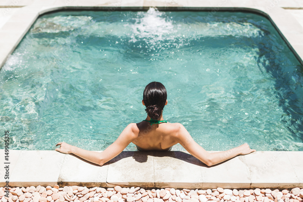 Woman in green swimsuit relaxing in outdoor jacuzzi with clean transparent turquoise water. Organic skin care in hot bath in luxury spa resort. - obrazy, fototapety, plakaty 