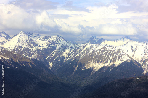 View from top of mountain peak. Mountain valley in blue haze and snowy mountain range. Ravine and cloudy sky. 