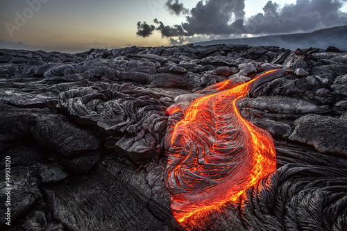 Red Orange vibrant Molten Lava flowing onto grey lavafield and glossy rocky land near hawaiian volcano with vog on background