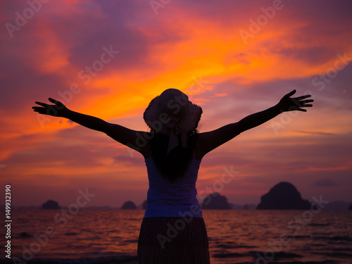 Silhouette of woman wearing hat with open arms under the sunrise near the sea © Siam