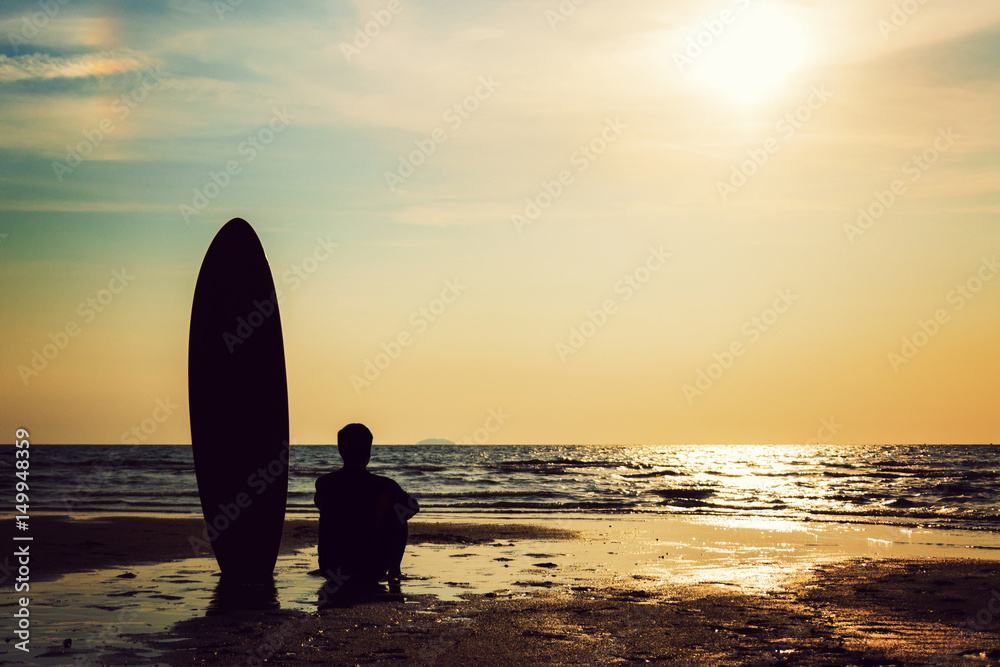 Silhouette of surf man sitting with a surfboard on the seashore beach at sunset time. Handsome Asia man model in his 20s.
