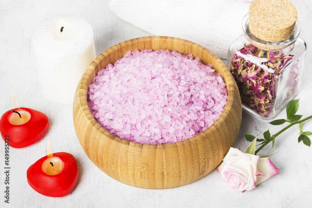 Bath salt with aroma of a rose in a wooden bowl, petals and a fresh pink rose, towels and candles on a white background