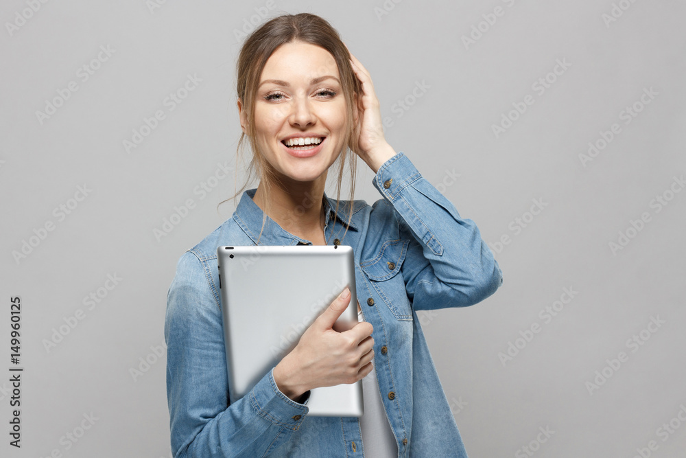 Indoor shot of cute European woman posing isolated on gray background with tablet PC in her hands, looking straight at camera, smiling and laughing cheerfully, looking confident and satisfied.