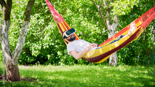 Young bearded man wearing virtual reality goggles relaxing in a garden hammock. Lifestyle VR fun and relax concept