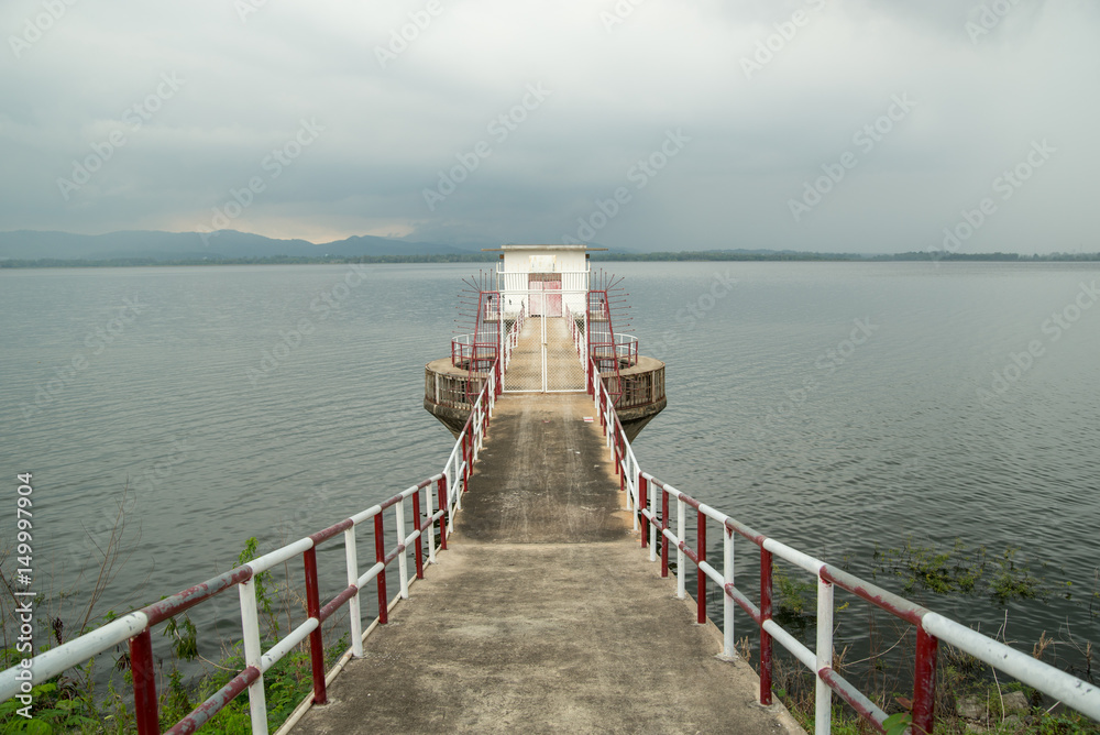 Wide-angle view of a concrete pier with white and red metal railings in the center, with a shed at the end. Bang Phra Reservoir, Chon Buri, Thailand.
