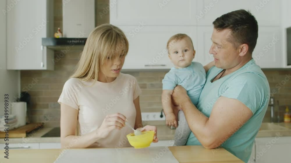 A young mother prepares to eat her 10 month old son. The father stands next to him and holds the child in his arms. Baby food, happy family with baby A young mother prepares to eat her 10 month old son. The father stands next to him and holds the child in his arms. Baby food, happy family with baby