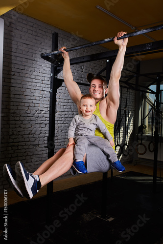 Young athlete father hangs on the horizontal bars and holds on his knees little son against brick wall.