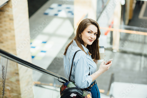sale, consumerism and people concept - beautiful young woman in striped shirt, blue jeans and bagpack holding mobile phone and disposable cup of coffee and rising on escalator in shopping center.