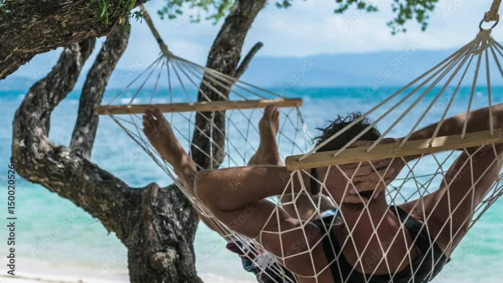 Man swinging relaxed in a hammock on the beach in front of the blue ocean. Hiding him from the sun in the shadow of a wizard trees.
