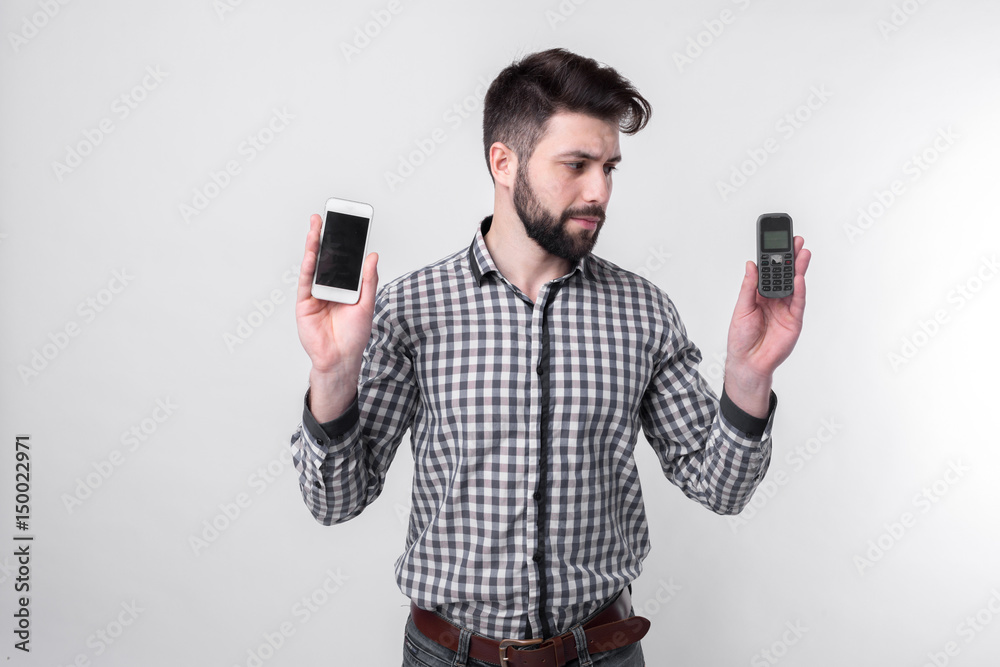 Bearded man isolated on a light background holding a modern smartphone and old cell phone with buttons Bearded man isolated on a light background holding a modern smartphone and old cell phone with buttons
