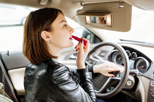 Businesswoman looking in rear view mirror and making up her lips with red lipstick in car