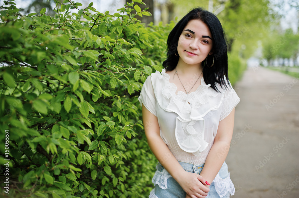 Portrait of sexy brunette girl on women's jeans shorts and white blouse against green spring bushes. Portrait of sexy brunette girl on women's jeans shorts and white blouse against green spring bushes.