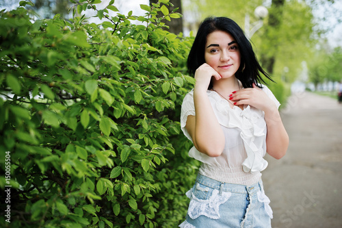 Portrait of sexy brunette girl on women's jeans shorts and white blouse against green spring bushes.