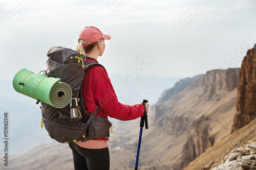 A slender girl in a cap with sticks for Nordic walking with a backpack and a folded rug for relaxation stands in the mountains against the backdrop of rocks and distant lands