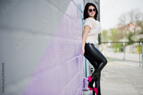 Portrait of brunette girl on women's leather pants and white blouse, sunglasses, against purple and gray wall.