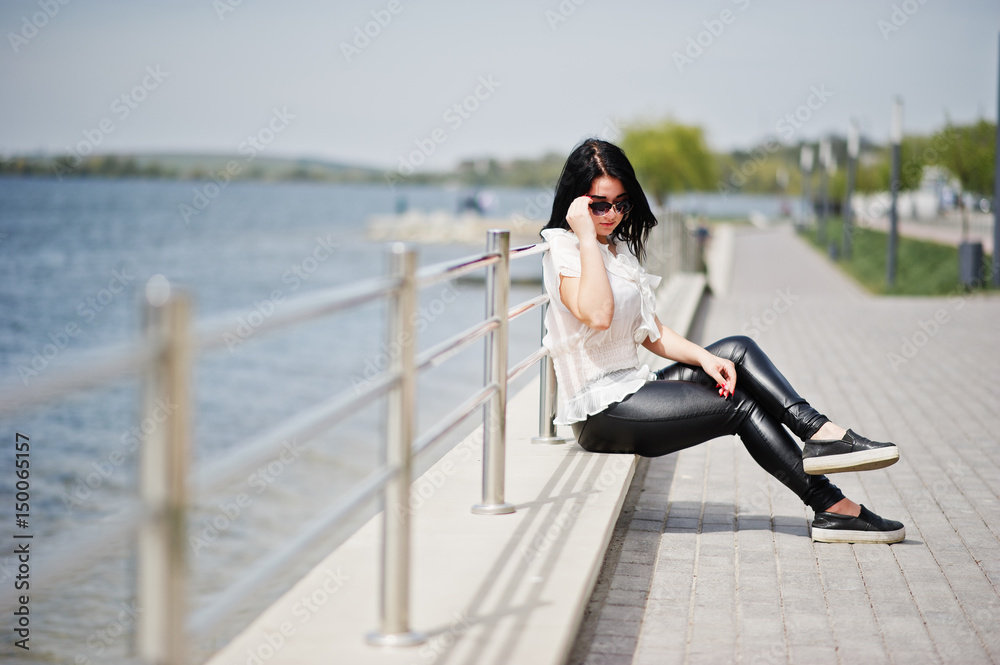 Portrait of  brunette girl on women's leather pants and white blouse, sunglasses, against iron railings at beach.