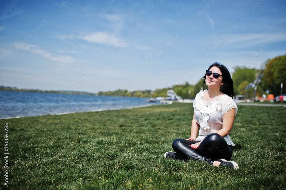 Brunette girl on women's leather pants and white blouse, sunglasses, sitting on geen grass against beach of lake. Brunette girl on women's leather pants and white blouse, sunglasses, sitting on geen grass against beach of lake.