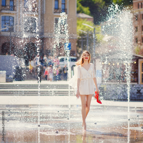 Young girl in a light dress is having fun in a fountain on the background of a sunny city