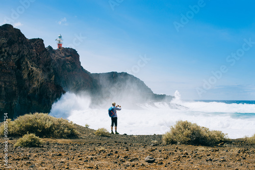A tourist with a backpack standing on the beach and photographs on phone large waves on the lighthouse background in the Canary Islands, Spain