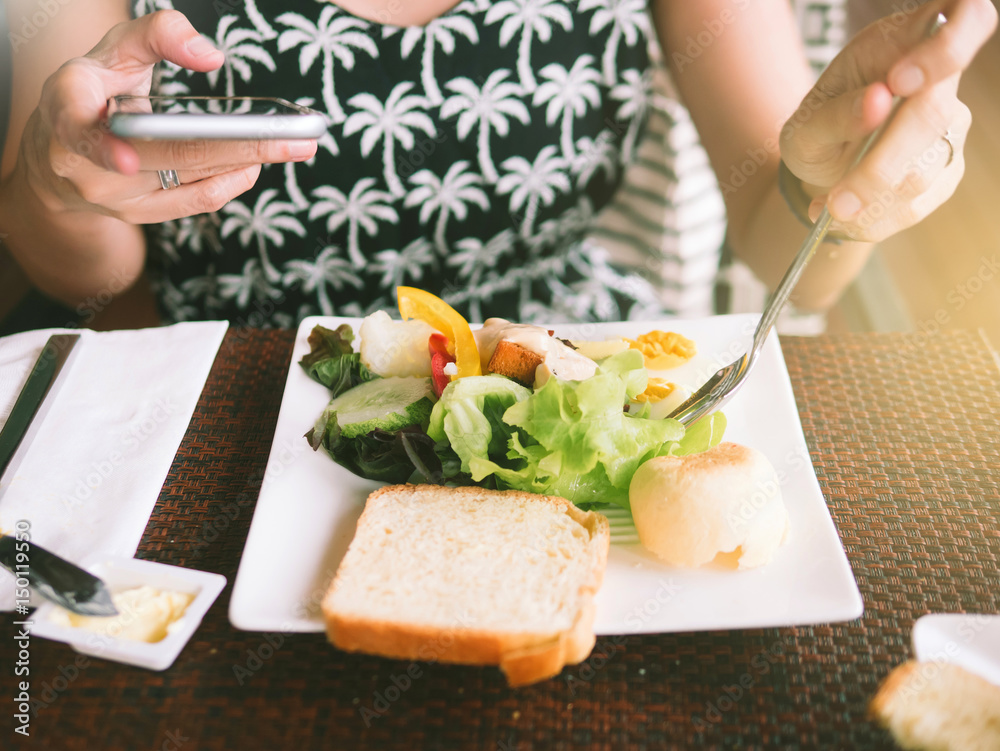 Closeup on a young woman's hands playing Mobile phone and eating breakfast. There is bread, butter, vegetable salad, Time For Breakfast