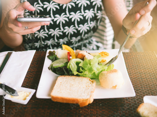 Obraz na płótnie Closeup on a young woman's hands playing Mobile phone and eating breakfast. There is bread, butter, vegetable salad, Time For Breakfast w salonie