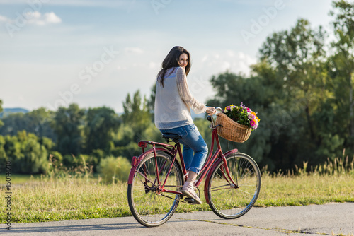 Young smiling woman rides a bicycle with a basket full of flowers in countryside