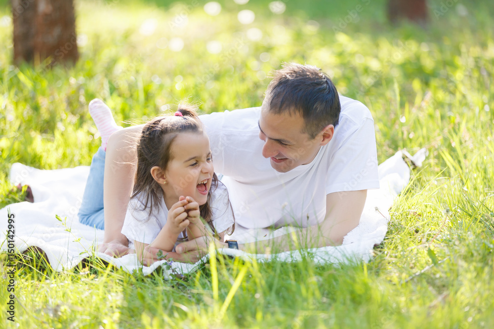 Young father and daughter lying on green grass. Happy daughter sitting on father back and smiling. looking up Young father and daughter lying on green grass. Happy daughter sitting on father back and smiling. looking up