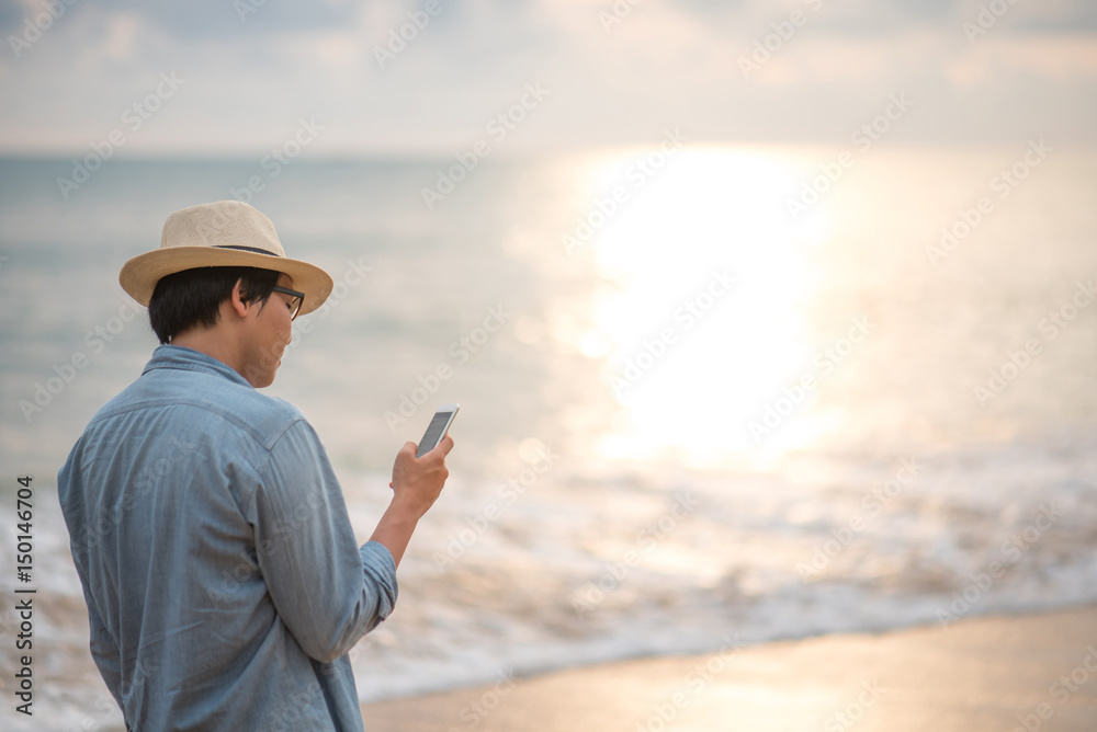 Young Asian man with jean shirt and hat holding smartphone and checking social media application on tropical beach at sunset, background for summer holiday vacation or global communication concepts