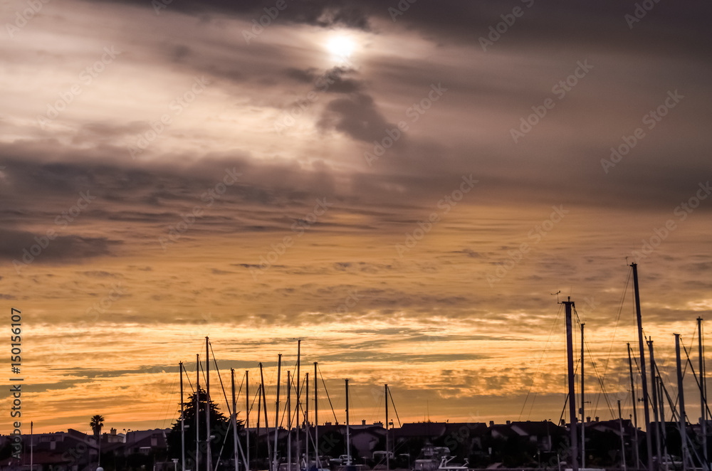 Silhouettes of boats on marina during cloudy yellow sunset in Oxnard, California