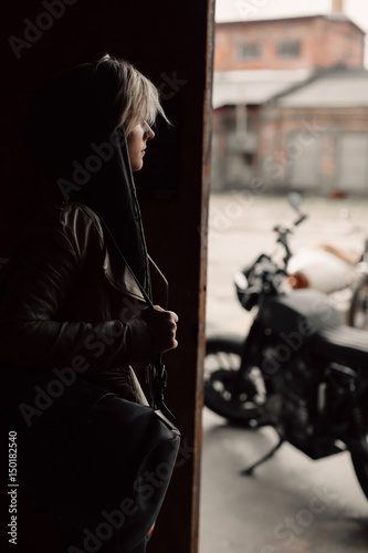 A young woman with a leather bag near the garage. A woman in a leather jacket. Motorcycle near the garage. Leather bag