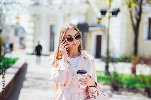 Upper class woman . Fashionable woman texting outdoors. Fashion woman in a sunglasses and pink jacket with coffee © Elena Kratovich