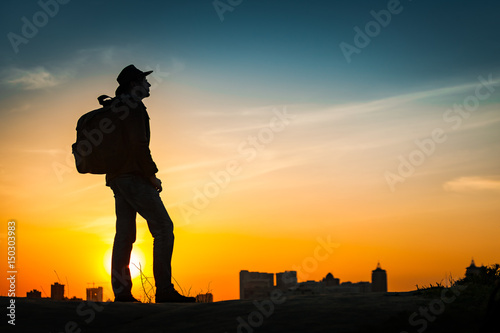 Traveler silhouette watching amazing sunset. Young casual man with backpack and cowboy hat standing alone on hill above evening cityscape. Lifestyle Travel Concept Outdoor Background. Kiev, Ukraine