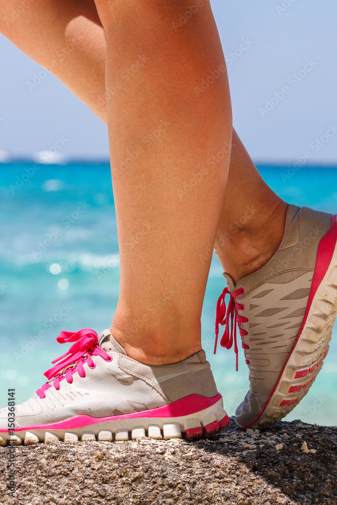Runner woman stands on a rock near the blue sea. Red laces gray sneakers. Closeup. Photo knee