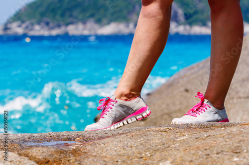 Runner woman stands on a rock near the blue sea. Red laces gray sneakers. Closeup. Photo knee