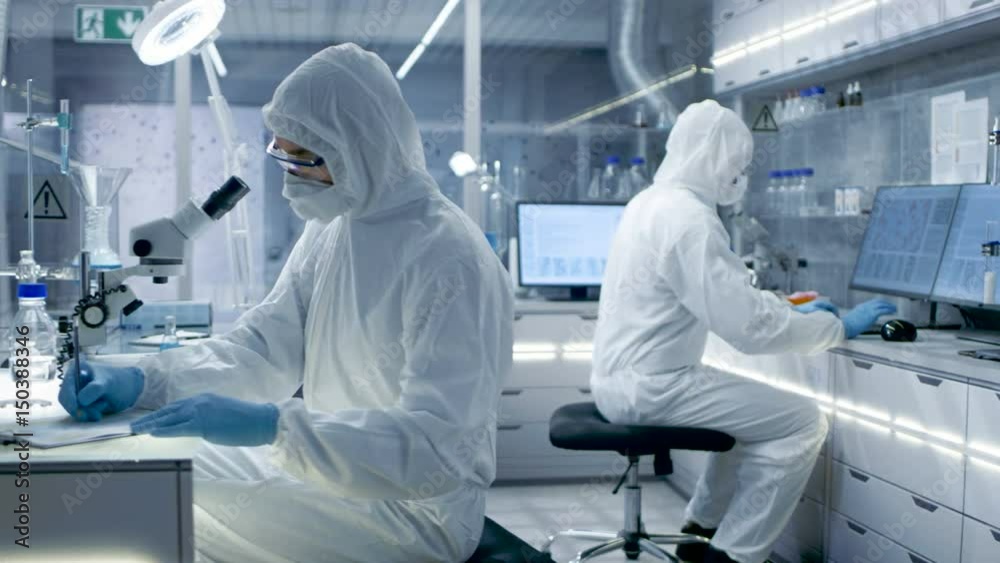 In a Secure High Level Laboratory Scientists in a Coverall Conducting a Research. Biologist Adjusts Samples in a  Petri Dish with Pincers and then Examines Them Under Microscope.
