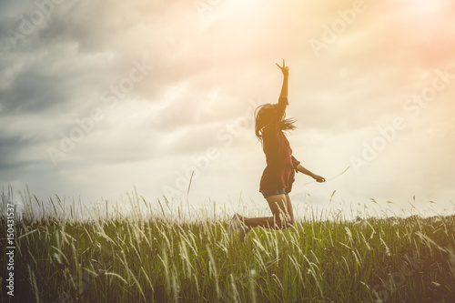 Fototapeta Naklejka Na Ścianę i Meble -  Woman jumping and raising up her hand and two finger over grass field on summer evening