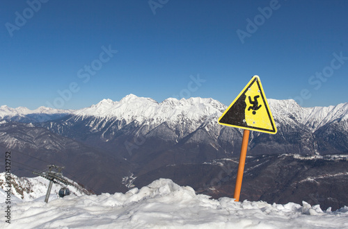 Горный пейзаж, знак предупреждающий об опасности падения, заснеженные вершины и горы с высоты Роза Пик. Хребет Аибга, Кавказ, Сочи, Россия