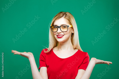 Portrait of Young Pretty Woman with Long Blond Hair, Eyeglasses and Red Top Holding Empty Space on Her Two Hands on Green Background in Studio.